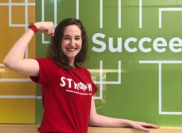 A woman in a red 'STRONG' shirt smiles confidently, flexing her arm in front of a green wall with the word 'Succeed' in bold white letters.