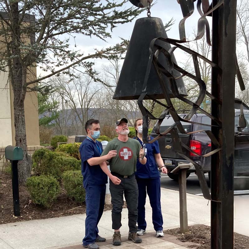 A man in a T-shirt is ringing a large metal bell outdoors, supported by two healthcare workers in scrubs and masks. The scene conveys a sense of achievement.