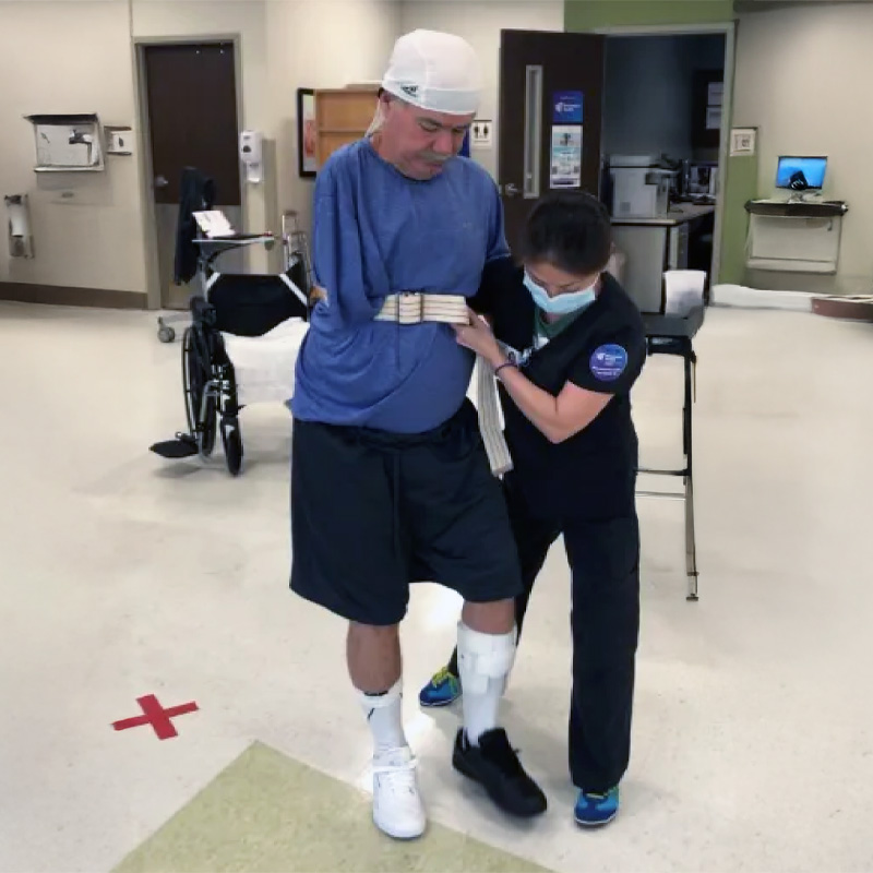 A person in a blue shirt and head bandage is learning to walk with support from a physical therapist in a hospital setting. A wheelchair is nearby.
