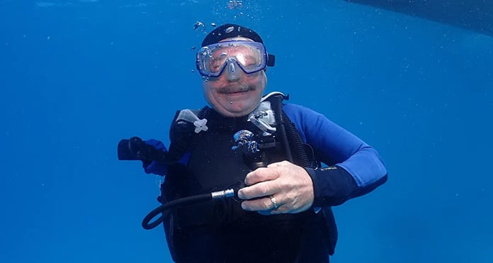 An underwater scene with a smiling scuba diver in full gear, including goggles, against a clear blue ocean backdrop. The tone is joyful and adventurous.