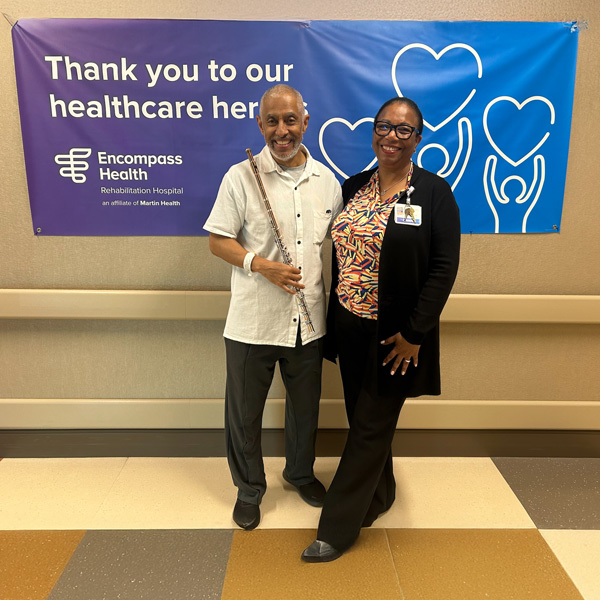 Nestor Torres holding a flute and posing with an Encompass Health CRRN
