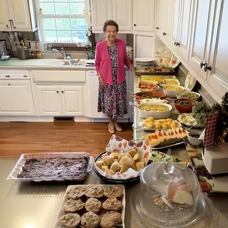 Elderly woman in bright kitchen smiling beside an extensive buffet with various casseroles, salads, desserts, and pastries, creating a warm, festive ambiance.