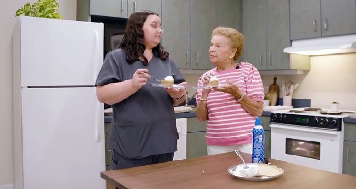Two women in a kitchen, one in scrubs and the other in a pink striped shirt, enjoy cake slices with whipped cream, sharing a warm moment.