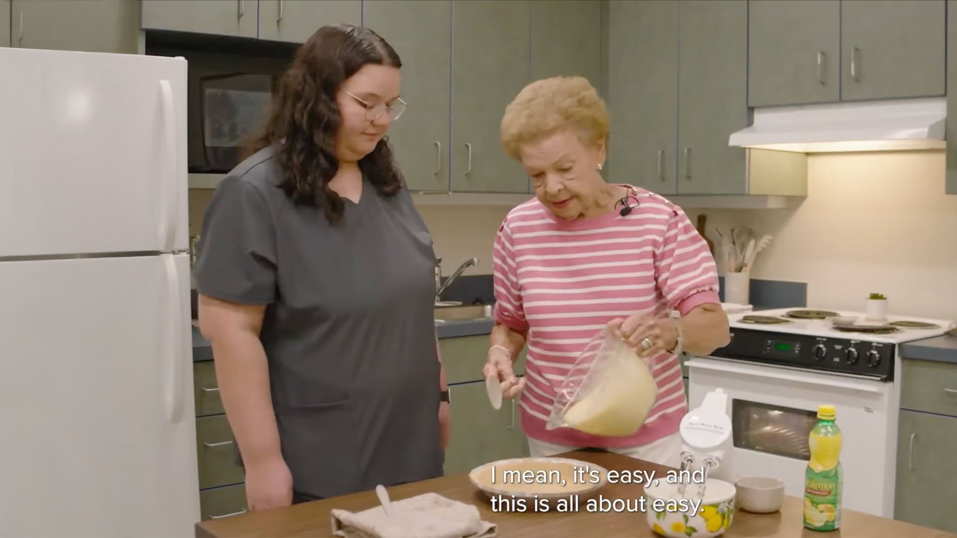 An elderly woman in a pink striped shirt pours batter into a dish while a younger woman in a gray top watches. They are in a kitchen, looking focused.