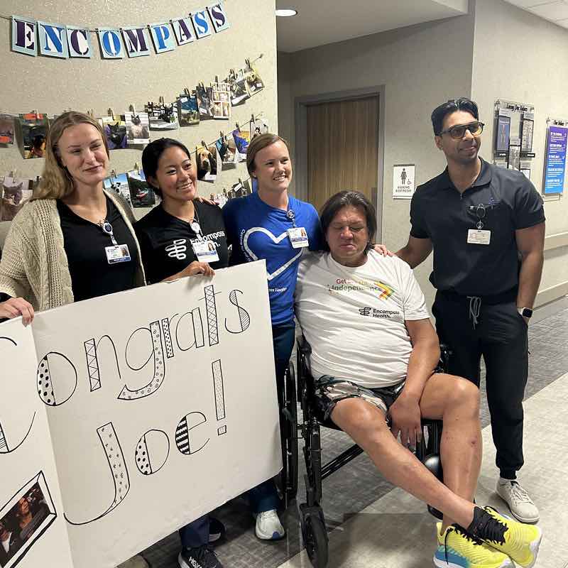 A group of four smiling healthcare workers stands with a man in a wheelchair holding a large 