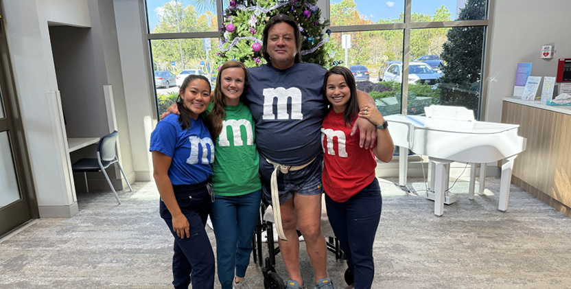Four people smiling and posing indoors, dressed in M&M-themed shirts in blue, green, and red. They stand together, conveying a joyful and friendly atmosphere.