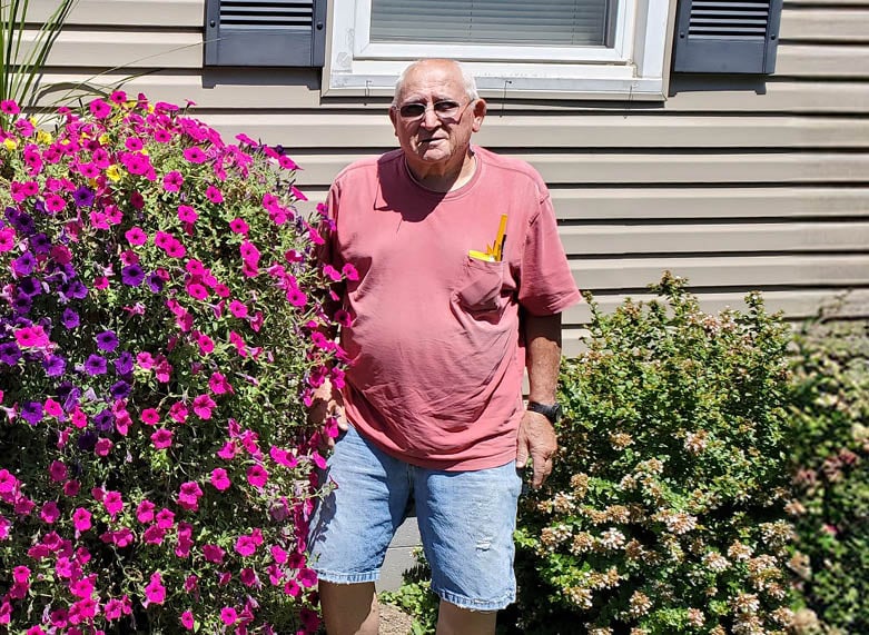 Howard K Jones in a pink shirt and denim shorts stands smiling near vibrant pink flowers and shrubbery outside a house on a sunny day.