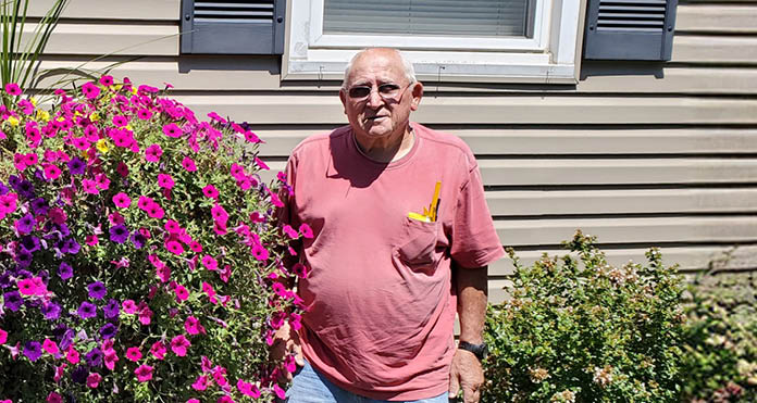 Howard K Jones in a pink shirt and denim shorts stands smiling near vibrant pink flowers and shrubbery outside a house on a sunny day.