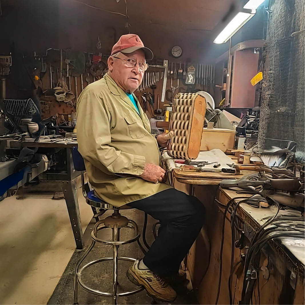 Howard K Jones in a workshop, wearing a red cap and khaki shirt, holding a wooden checkerboard. Surrounded by tools, the room has a rustic charm.