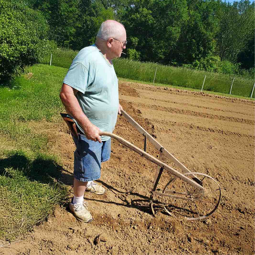 Howard K Jones stands in a garden, using a manual plow on freshly tilled earth. He wears a light blue shirt and denim shorts. The background is green and lush.