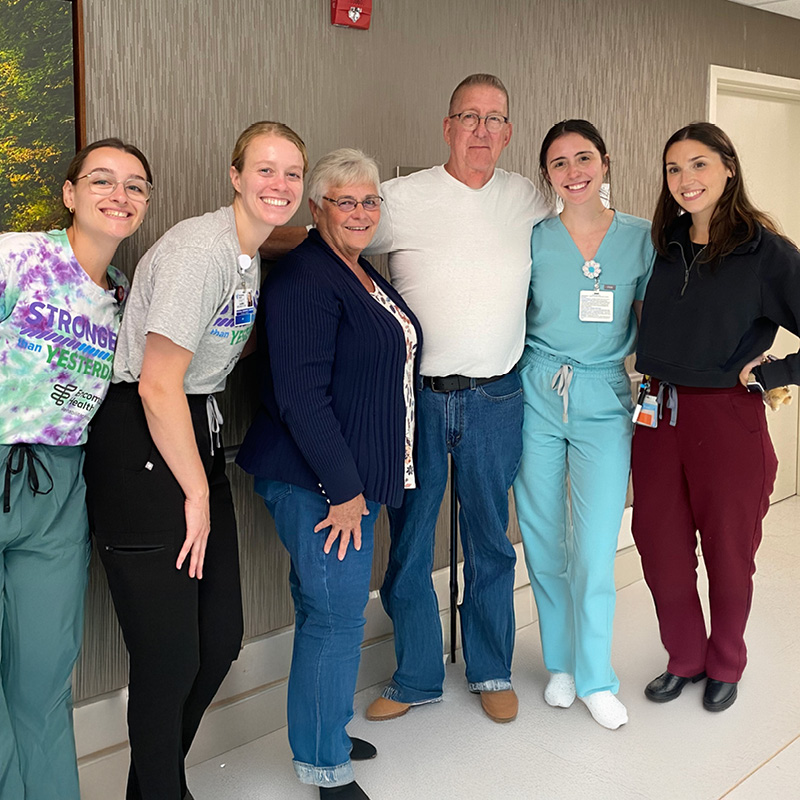 A group of six people stand in a hallway, smiling warmly. The man and woman in the center are flanked by four hospital staff in casual uniforms, showing camaraderie.