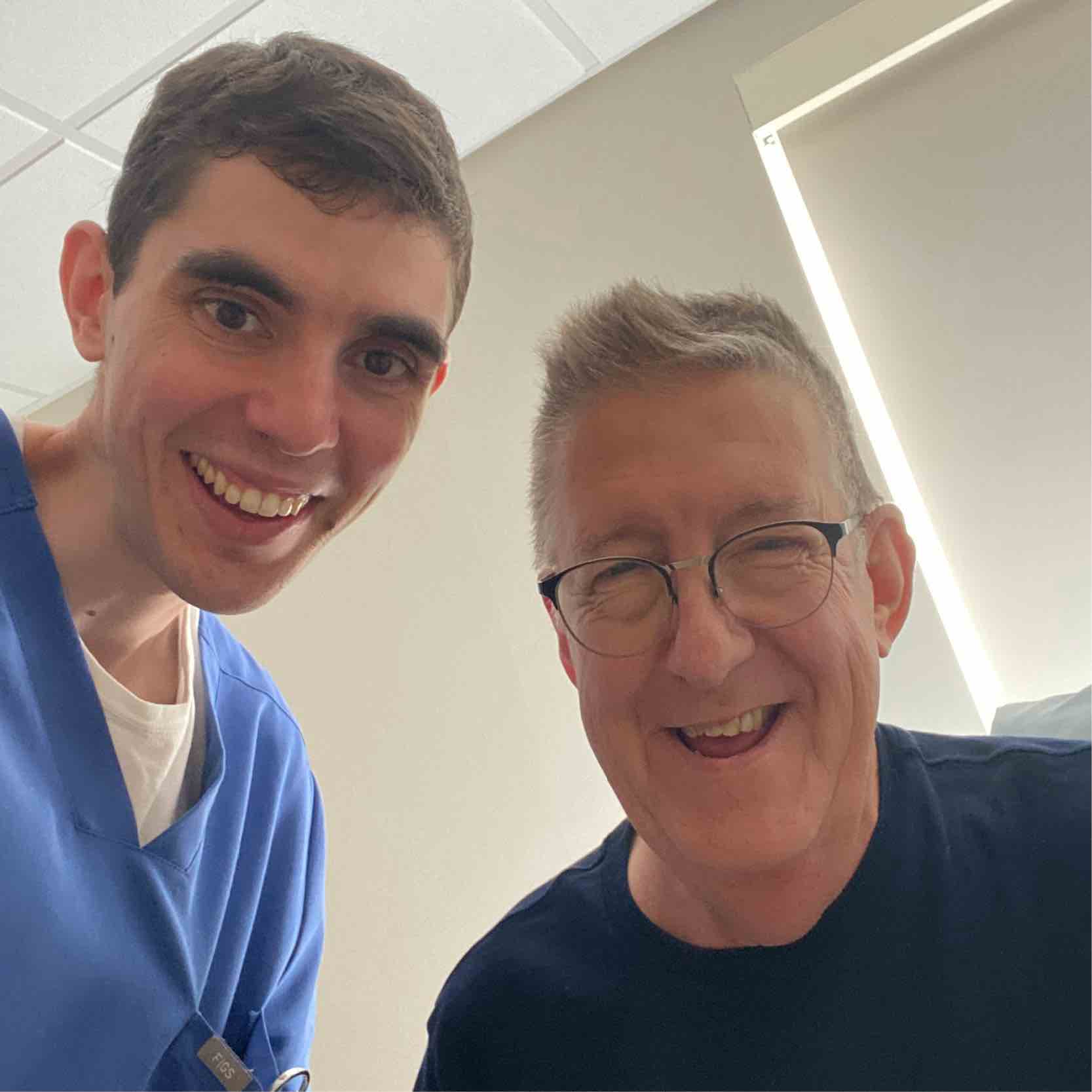 Two men smiling warmly, with one wearing blue scrubs, suggesting a healthcare setting. Bright, friendly atmosphere with soft lighting.
