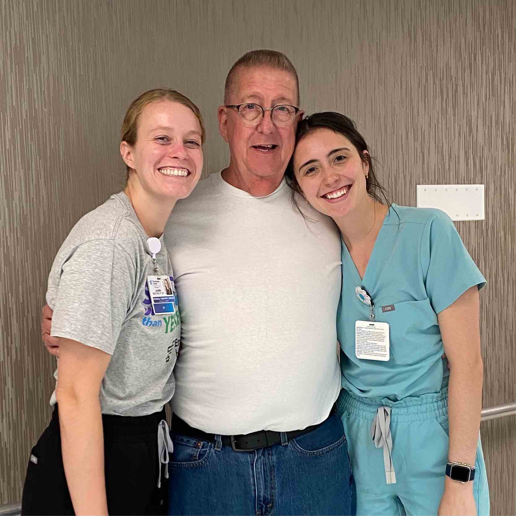 An older man in a white shirt stands between two smiling nurses, one in scrubs. They are in a clinical setting, exuding warmth and camaraderie.