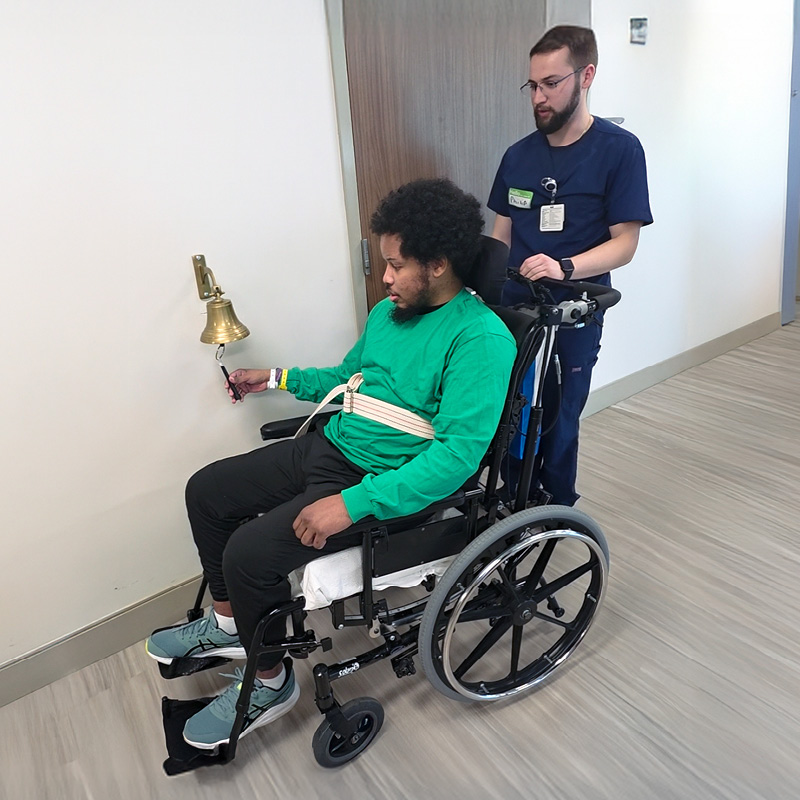 A man in a wheelchair, wearing a green shirt, rings a bell in a hallway. He is assisted by a healthcare worker in blue scrubs behind him. The atmosphere is uplifting.
