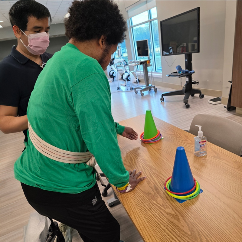 A person wearing a green shirt and a support belt engages in physical therapy, reaching for colorful cones on a wooden table. Another person assists nearby. The room is bright, with exercise equipment and a flat-screen TV visible in the background, conveying a rehabilitative setting.