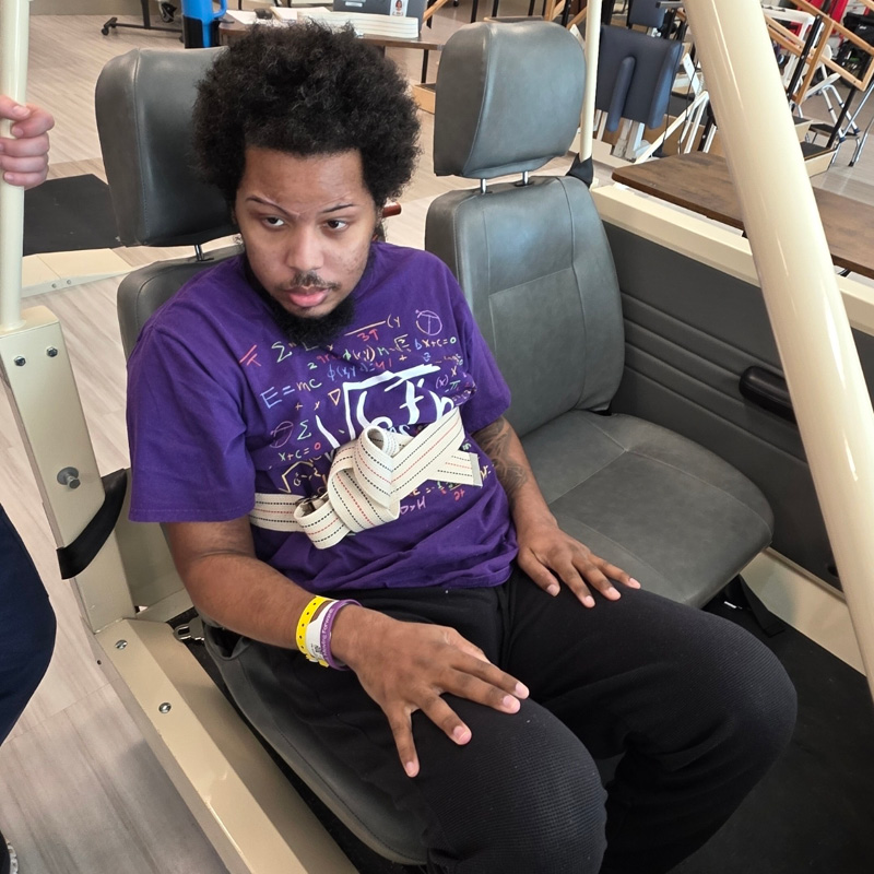 A man with an afro, wearing a purple shirt with white patterns, sits strapped into a gray seat. His expression is focused. He wears wristbands.