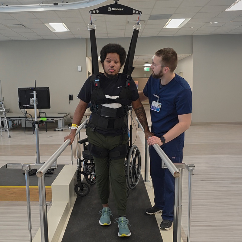 Man in Vector harness uses rehabilitation equipment; assisted by a therapist in a medical setting. Focused expressions, supportive atmosphere.