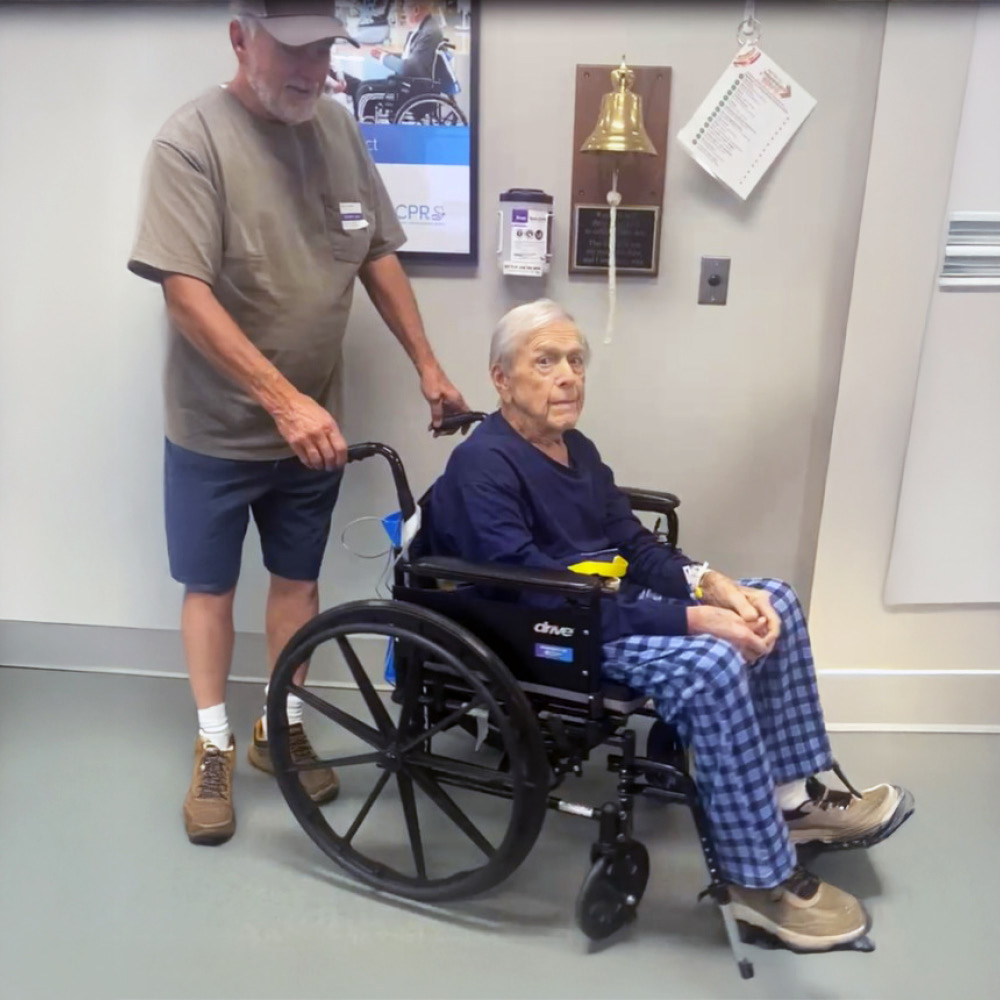 A senior man in a wheelchair is pushed by another man in a casual outfit. A bell is mounted on the wall behind them, suggesting a hospital or clinic setting.