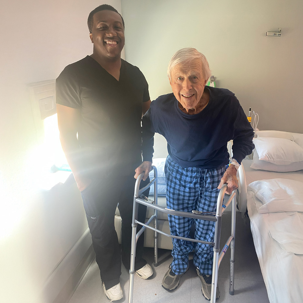 A smiling healthcare worker stands beside an elderly man using a walker in a bright room with a bed. The mood is cheerful and supportive.