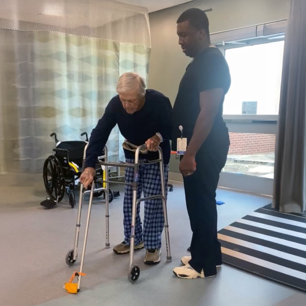 An elderly person uses a walker in a rehab room, guided by a caregiver. A wheelchair and wide window add to the supportive atmosphere.
