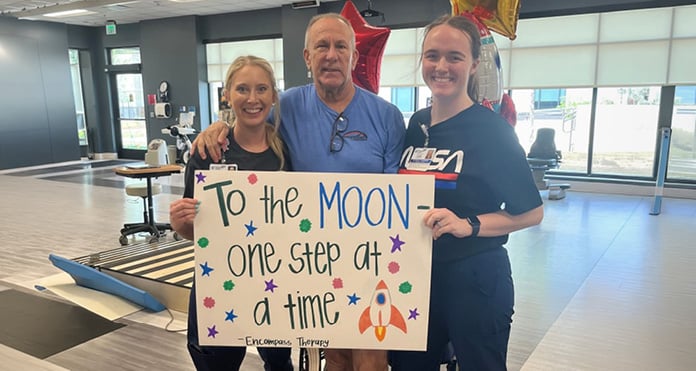 Three people pose in a rehabilitation gym holding a sign that reads, To the MOON one step at a time, celebrating a therapy milestone.