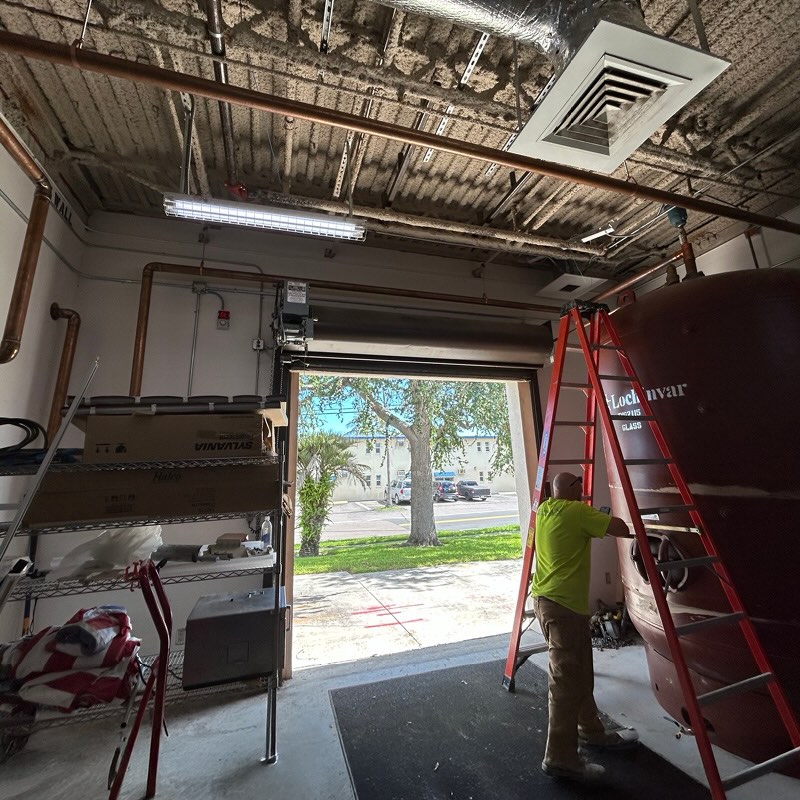 Worker in a neon shirt on a red ladder adjusts equipment in a garage. Open door reveals a sunny street with trees and parked cars outside.
