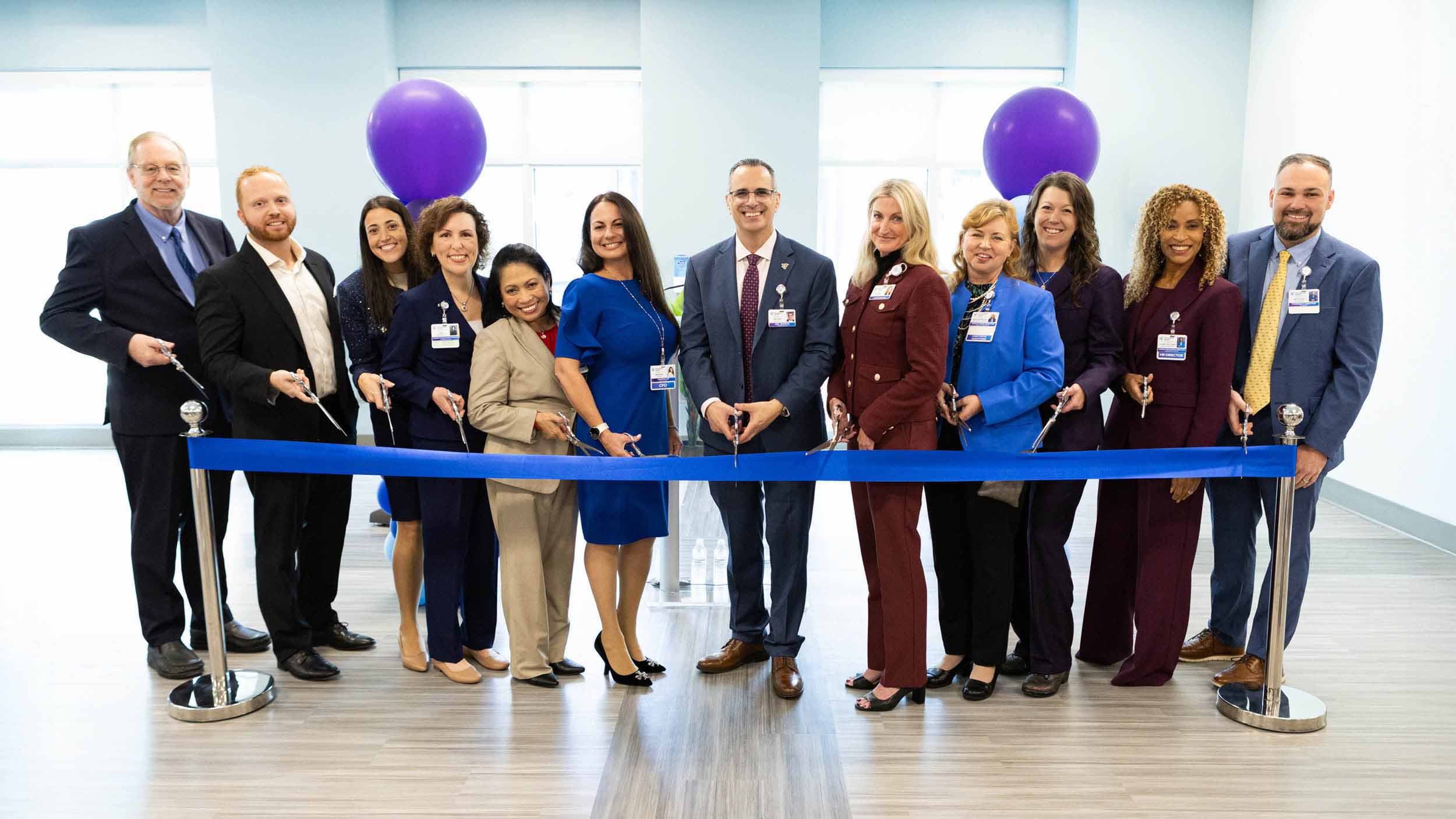 A group of twelve people stand smiling at a ribbon-cutting ceremony. They hold scissors behind a blue ribbon in a bright room with purple balloons.
