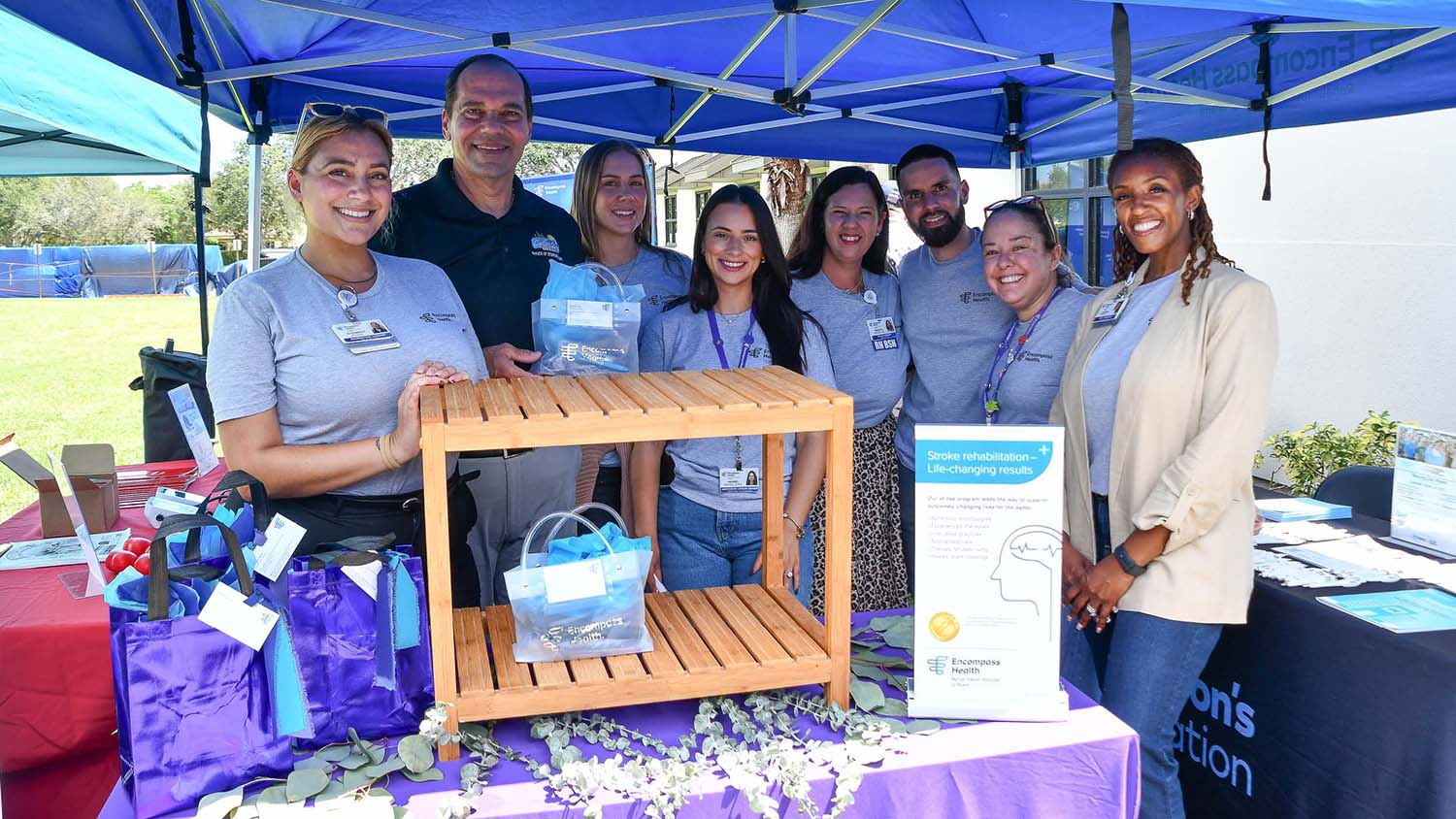 A group of people in branded shirts stand smiling under a blue tent at an outdoor event. A wooden display rack and informational sign are in front of them.