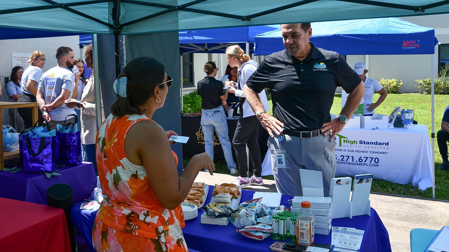 A woman in a floral dress talks to a man at an outdoor booth under a tent, with snacks and brochures displayed. People chat at nearby booths. The mood is lively and engaged.