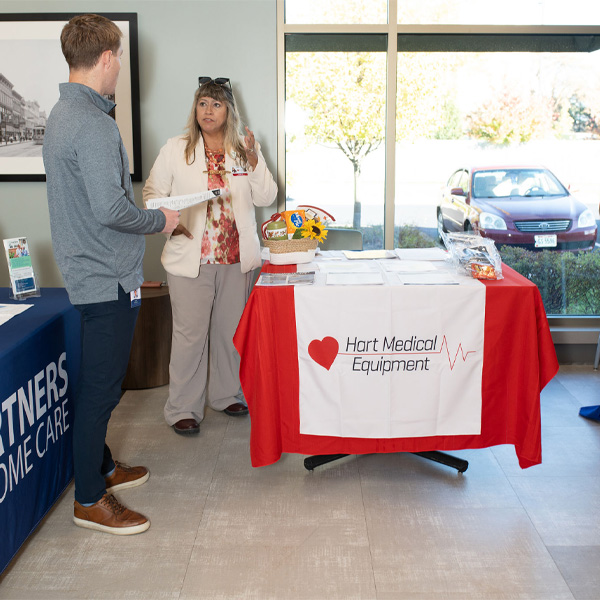 A woman and man at the Encompass Health Toledo open house event converse at a Hart Medical Equipment booth with a red tablecloth displaying pamphlets and a basket.