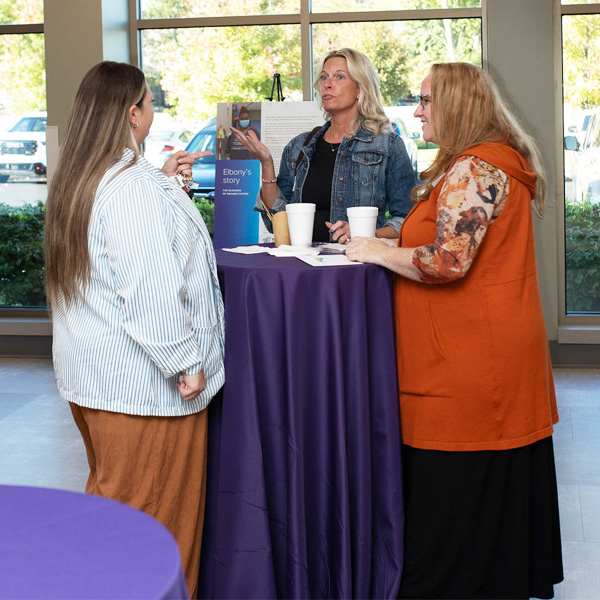 Three women at the Encompass Health Toledo open house event converse around a high table with a purple tablecloth, holding drinks.