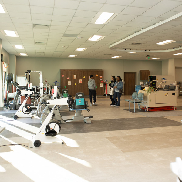 The Encompass Health Toledo therapy gym with exercise bikes and equipment is well-lit by overhead lights and daylight. Three people converse warmly.