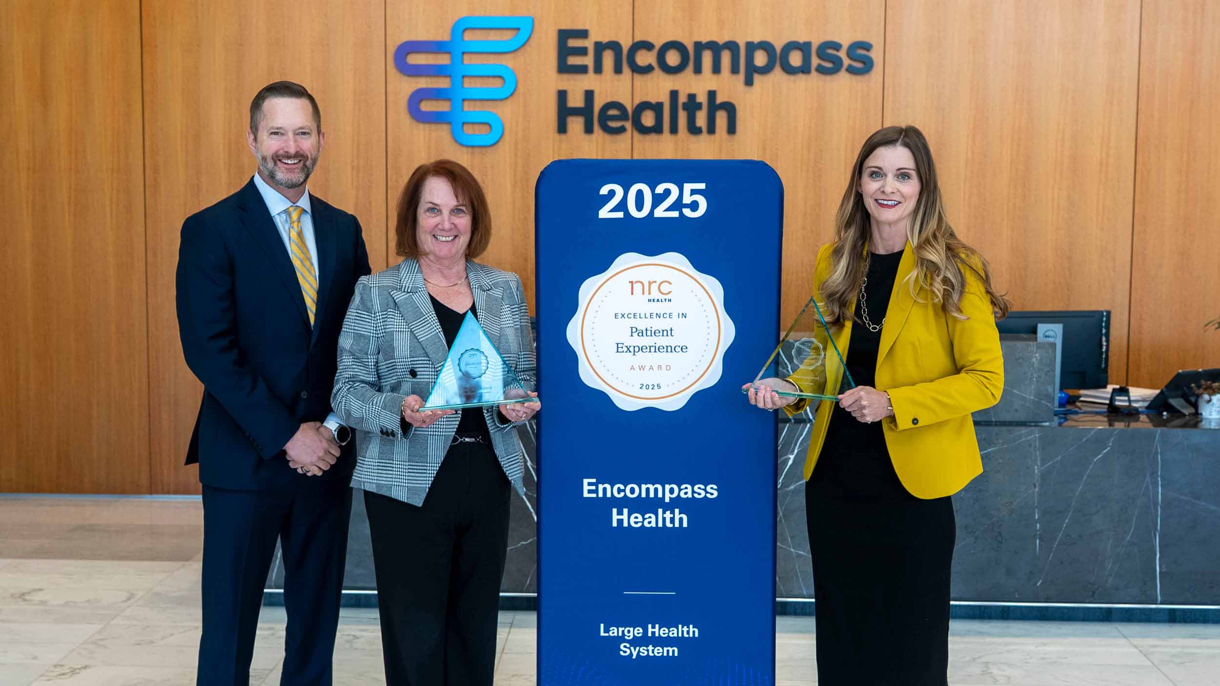Three people stand smiling in front of an Encompass Health sign. They hold awards for Excellence in Patient Experience, conveying a sense of achievement.