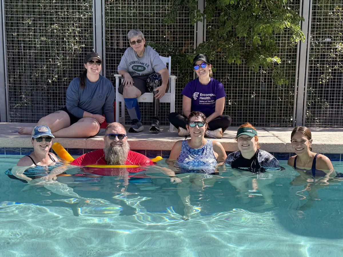 A group of seven adults at a pool, with four in the water and three seated on the deck. They are smiling under a sunny sky, exuding a joyful vibe.
