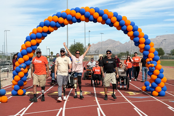 A group of people with prosthetic legs and in wheelchairs celebrate under an arch of blue and orange balloons on a track, showing unity and joy.