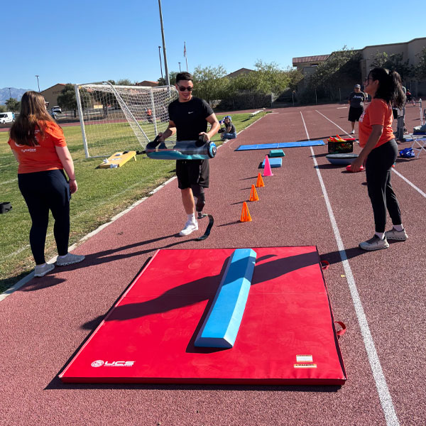 Three people are on a track beside a soccer field, engaging in an obstacle course. A man carries a mat near a red pad and orange cones. It's a sunny day.
