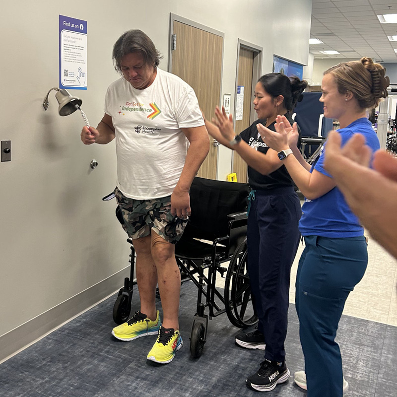 A man in a white T-shirt and shorts rings a bell while standing beside a wheelchair. Two people in uniforms cheer and clap, conveying a celebratory tone.