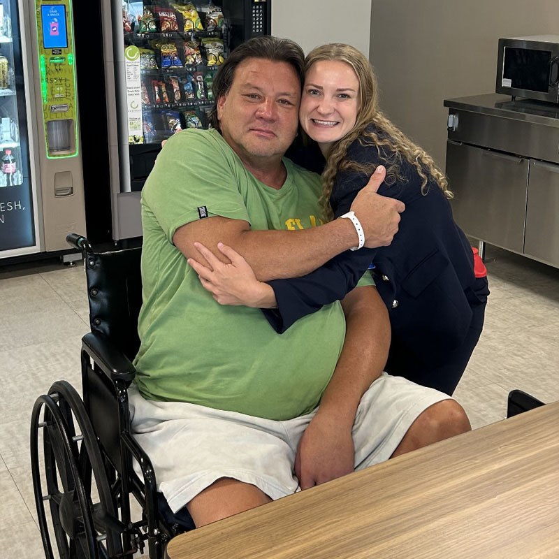 A man in a green shirt, sitting in a wheelchair, is hugged by a smiling woman in a blue outfit. They are in a room with vending machines.