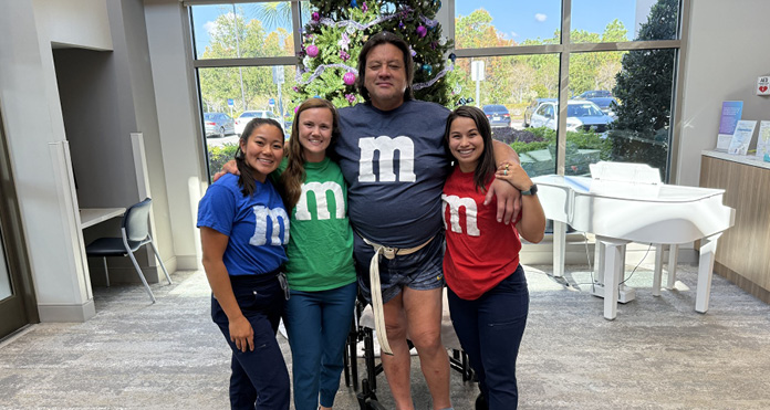 Four people smiling and posing indoors, dressed in M&M-themed shirts in blue, green, and red. They stand together, conveying a joyful and friendly atmosphere.