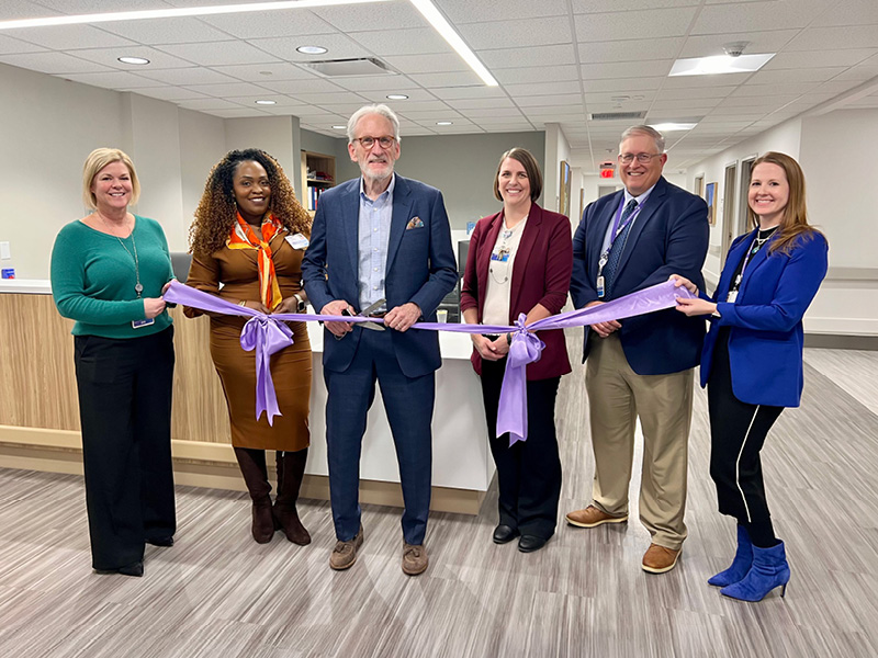 A group of six employees at Encompass Health Rehabilitation Hospital of New England stand in the expanded ReACT unit smiling and holding a purple ribbon for a ribbon-cutting ceremony.