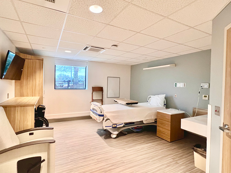 A modern hospital room with a single bed, white walls, and wood accents. A large window brings in natural light, creating a calm and welcoming atmosphere.