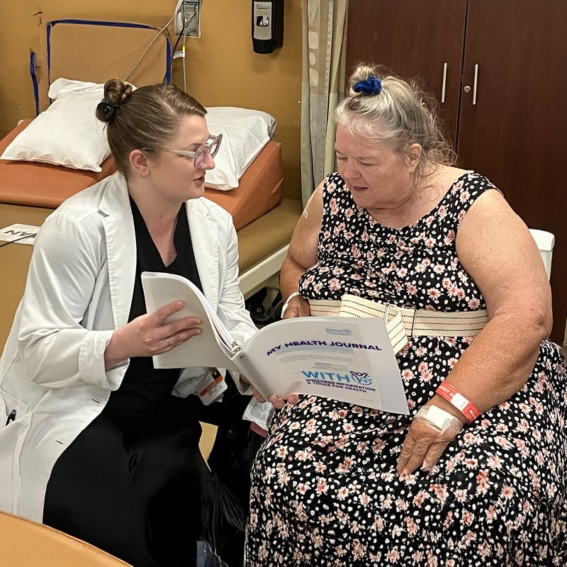 OT Intern Ashley Brewer discusses a WITH book with an older woman in a floral dress. They sit together in a hospital room, focusing on the document with a supportive, engaged expression.