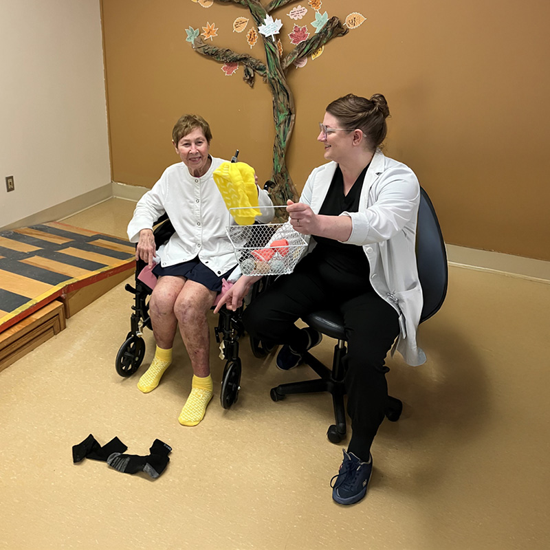 A smiling elderly woman in a wheelchair, wearing bright yellow socks, engages with OT intern Ashley Brewer holding a basket of therapy items, set in a warm room with a decorative paper tree on the wall.