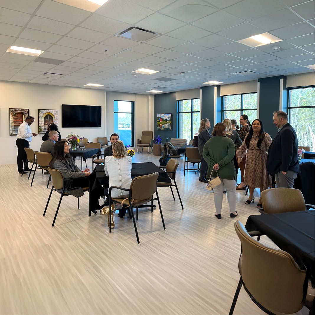 A spacious dayroom with a group of people engaging in conversation around tables. Large windows line the walls, creating a bright, welcoming atmosphere.
