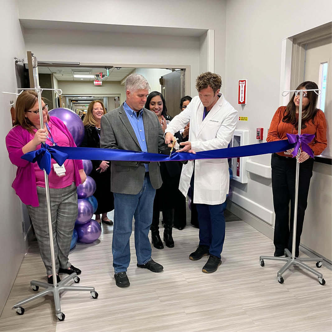 Doctor in a white coat cuts a blue ribbon at a ceremonial opening, flanked by smiling colleagues and purple balloons, in a hospital corridor.