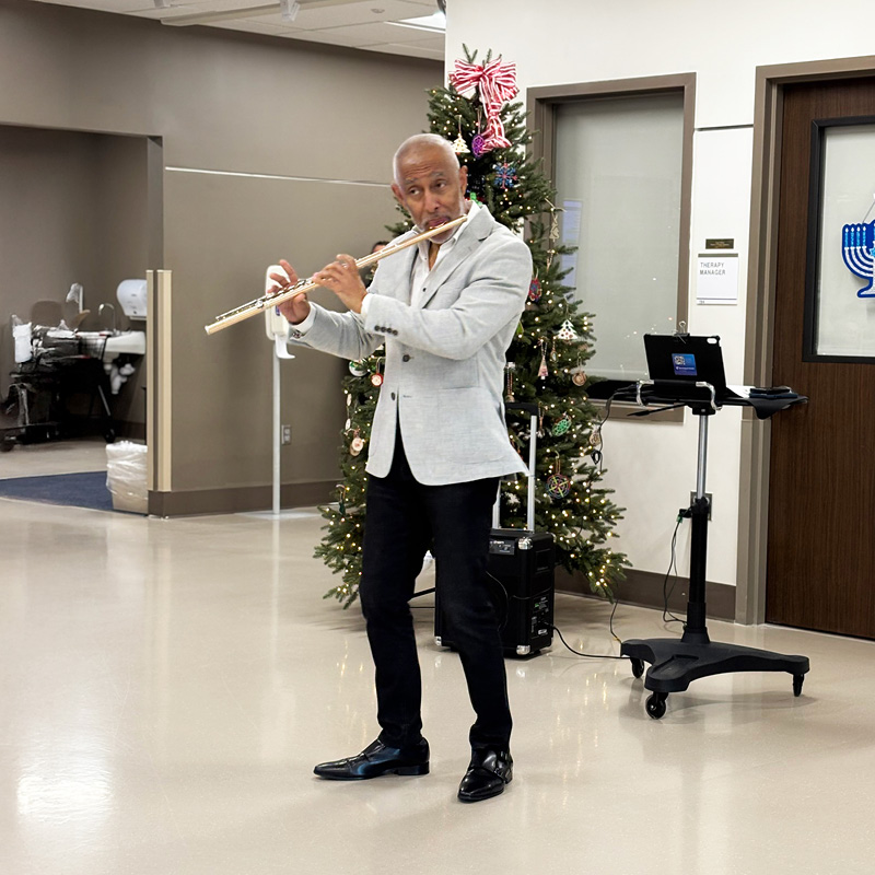 A man in a light blazer plays the flute in a festive setting next to a decorated Christmas tree. The atmosphere is warm and cheerful.