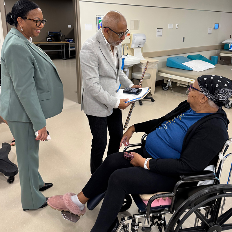 A professional and a flute player engage warmly with a patient in a wheelchair in a clinical setting. The atmosphere is friendly and supportive.