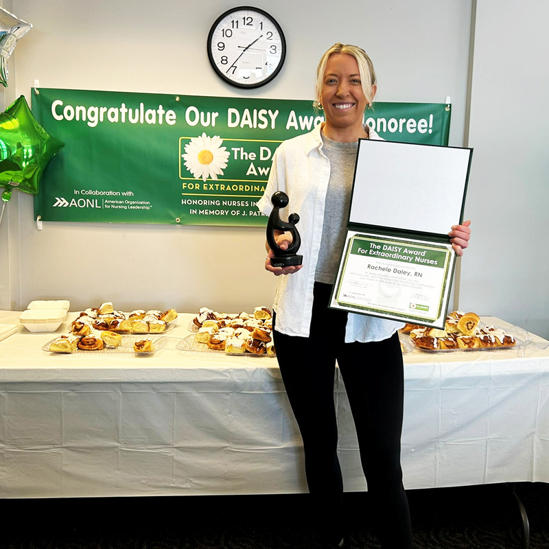 Smiling woman holds a DAISY Award and certificate in front of a congratulatory banner. A table with pastries and balloons is in the background.