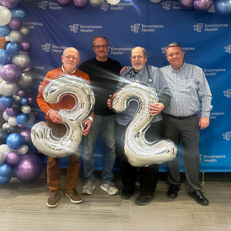 Four men stand in front of an Encompass Health backdrop holding large silver balloons shaped like the numbers 3 and 2, with balloon decorations on the left side.