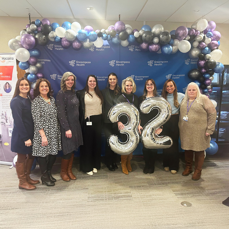 Nine women stand in front of a balloon arch and Encompass Health backdrop, with two women holding large silver balloons shaped as the number 32.